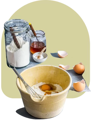 Flour, cracked eggs, and a whisk on a bowl. Egg shells and jars of some powders and condiments behind it.